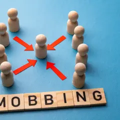 Drewniane ludziki otaczają jednego, a czerwone strzałki wskazują na niego. Słowo "mobbing" ułożone z liter.