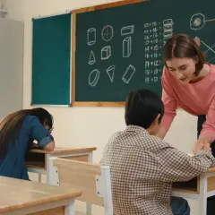 Nauczycielka pomaga uczniowi przy biurku, gdy jest wystawienie ocen. Na tablicy matematyka i owoce.