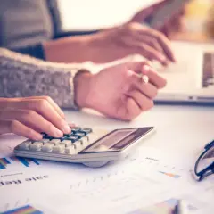 Dwie osoby pracują przy biurku, jedna wpisuje dane na kalkulatorze, druga na laptopie. Na stole leżą raporty, okulary i kubek. Obliczanie kilometrówki.