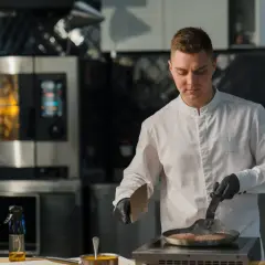 Młody kucharz w białej kurtce i czarnych rękawiczkach smaży mięso na patelni. To dowód jego kwalifikacji w zawodzie kucharz.