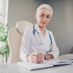Lekarka w białym kitlu z niebieskim stetoskopem pisze w notatniku, być może wystawia zwolnienie lekarskie wstecz.