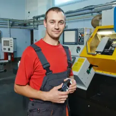 Uśmiechnięty tokarz w warsztacie, trzymający przyrząd pomiarowy, obok nowoczesnej maszyny CNC.