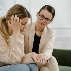 Kobieta w beżowej marynarce pociesza zapłakaną koleżankę, która zmaga się z problemami, być może związanymi z siłą wyższą i kodeksem pracy.