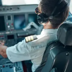 Pilot samolotu w słuchawkach i okularach przeciwsłonecznych, z ręką na sterach, w kabinie pełnej ekranów.