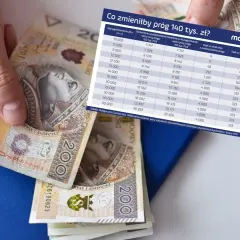 Dłoń trzyma banknot 200 zł. Obok leży niebieski notatnik i długopis. Na notatniku znajduje się tabela z danymi o wynagrodzeniach i wpływie przekroczenia progu podatkowego.