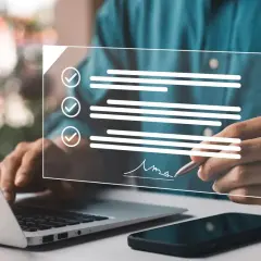 Dłoń podpisująca elektronicznie dokument na ekranie, obok laptop i telefon.