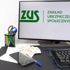 Biurko z monitorem wyświetlającym logo ZUS, pieniędzmi, telefonem i artykułami biurowymi.