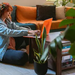 Kobieta z rudymi włosami pracuje na laptopie, siedząc na podłodze obok kanapy. To idealny przykład elastycznej **pracy hybrydowej** w domowym zaciszu.