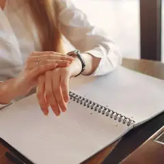 Kobieta w białej koszuli sprawdza zegarek, pracując po godzinach. Na biurku leży otwarty notatnik i laptop.