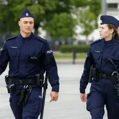 Dwóch policjantów w granatowych mundurach, z widocznymi stopniami w policji, patroluje teren.