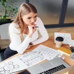 Młoda kobieta analizuje dokumenty, zastanawiając się, ile zarabia projektant mody. Na biurku laptop, szkice i kawa.