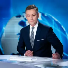 Elegancki konferansjer w garniturze stoi przy biurku na tle niebieskiej grafiki z globusem.
