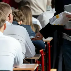 Uczniowie siedzą w ławkach, czekając na egzamin zawodowy sprzedawca. Nauczyciel rozdaje arkusze z zadaniami.