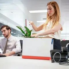 Kobieta pakuje rzeczy do kartonu, mężczyzna siedzi przy biurku.