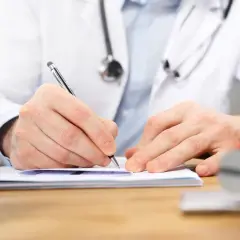 Lekarz w białym kitlu pisze długopisem na dokumentach, obok pieczątki.