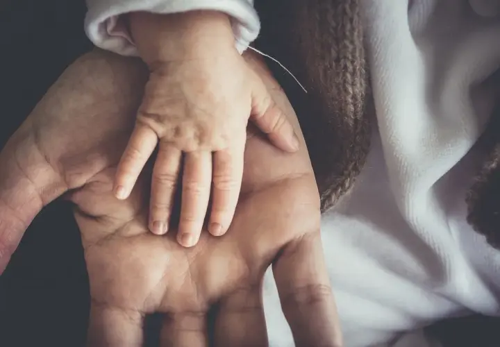 Dłoń dorosłego obejmuje dłoń niemowlęcia. To symbol opieki na dziecko do ilu lat, która trwa przez całe życie.