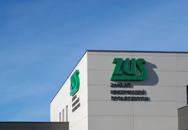 Budynek ZUS z zielonym logo i napisem "Zakład Ubezpieczeń Społecznych" na tle błękitnego nieba.