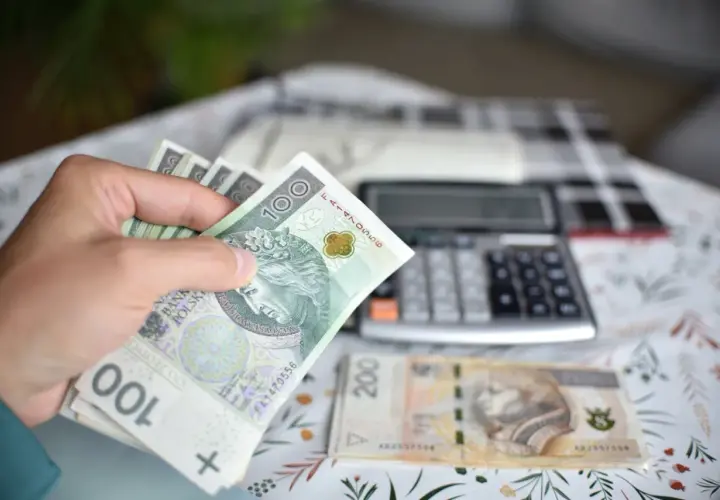 Ręka trzyma banknoty 100 zł, obok leży 200 zł i kalkulator. Obliczenia ekwiwalentu za urlop.