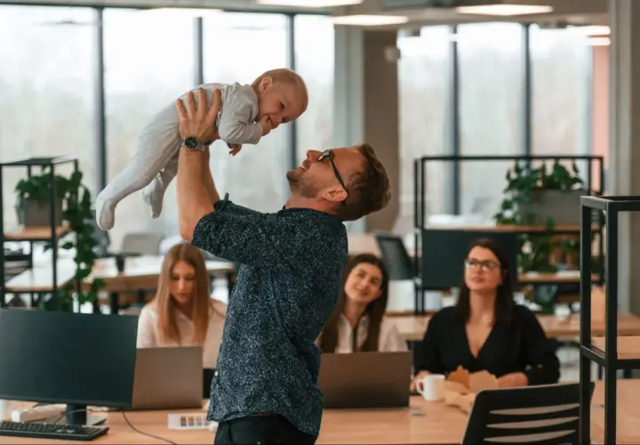 Mężczyzna w okularach podnosi dziecko do góry w biurze, w tle pracują kobiety.