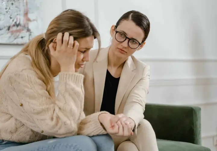Kobieta w beżowej marynarce pociesza zapłakaną koleżankę, która zmaga się z problemami, być może związanymi z siłą wyższą i kodeksem pracy.