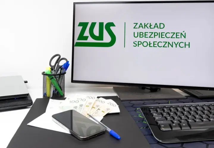 Biurko z monitorem wyświetlającym logo ZUS, pieniędzmi, telefonem i artykułami biurowymi.