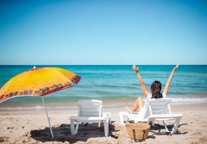 Kobieta cieszy się urlopem wypoczynkowym na plaży, z rękami w górze. Obok leżaki i parasol.