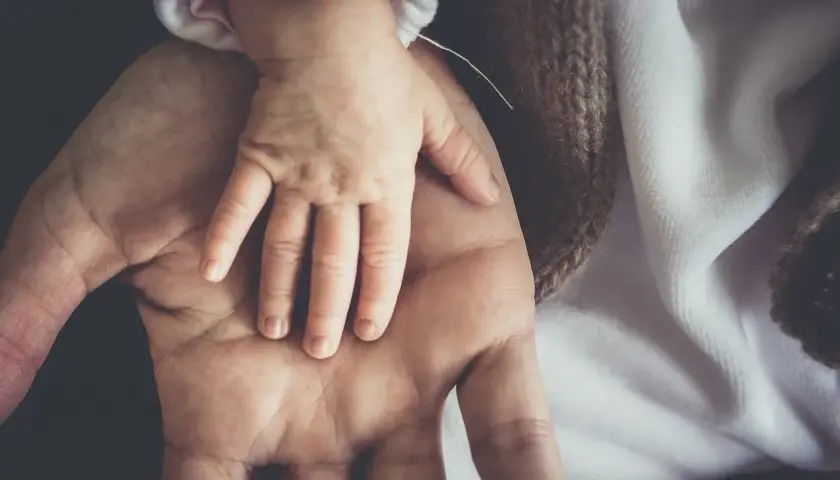 Dłoń dorosłego obejmuje dłoń niemowlęcia. To symbol opieki na dziecko do ilu lat, która trwa przez całe życie.