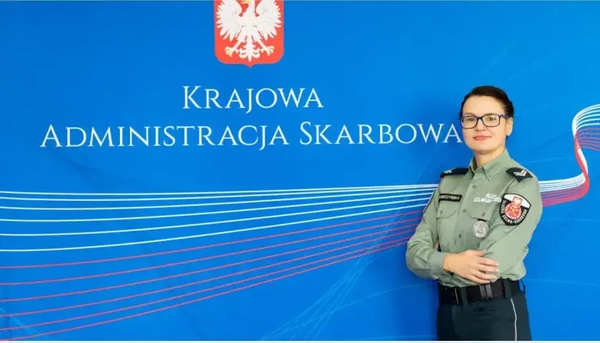Kobieta w mundurze służby celno-skarbowej stoi przed niebieskim tłem z napisem "Krajowa Administracja Skarbowa".