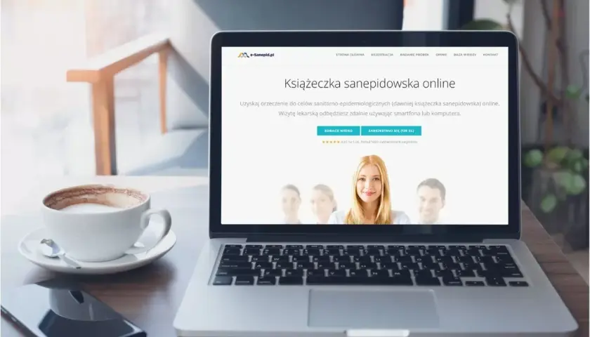 Laptop z kawą i telefonem. Na ekranie widnieje strona internetowa z ofertą "Książeczka sanepidowska online".