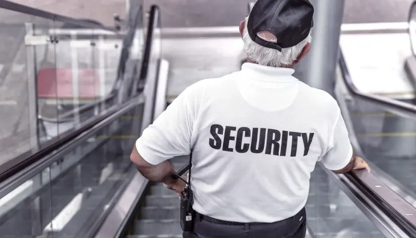 Pracownik ochrony w białej koszulce z napisem "SECURITY" stoi na ruchomych schodach, trzymając krótkofalówkę.