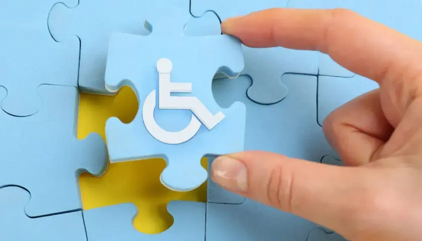 Dłoń układa puzzla z symbolem osoby na wózku inwalidzkim. Pfron druki do pobrania.
