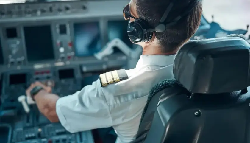 Pilot samolotu w słuchawkach i okularach przeciwsłonecznych, z ręką na sterach, w kabinie pełnej ekranów.