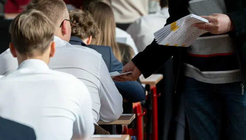 Uczniowie siedzą w ławkach, czekając na egzamin zawodowy sprzedawca. Nauczyciel rozdaje arkusze z zadaniami.