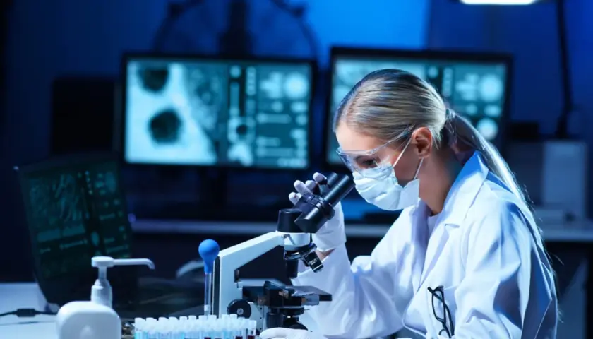 Diagnosta laboratoryjny w okularach i masce analizuje próbki pod mikroskopem. Na ekranach komputera widoczne są wyniki badań.