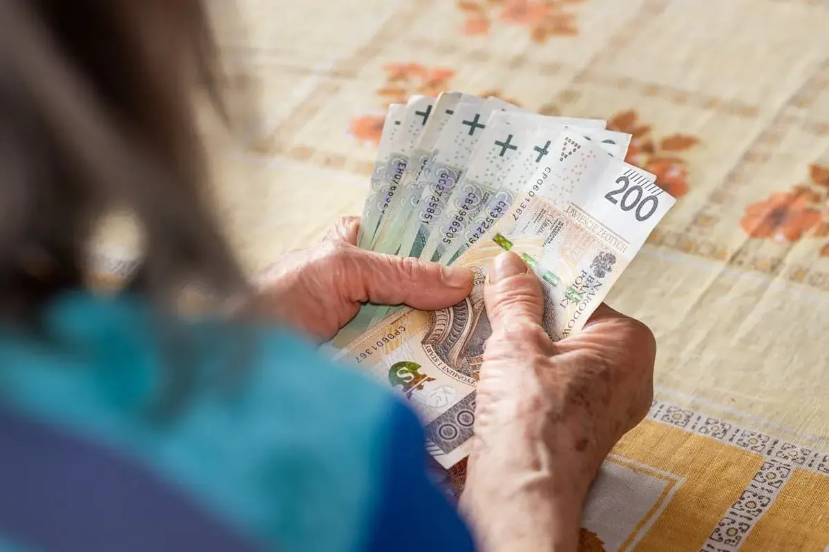 Dłonie starszej osoby liczą banknoty. W tle widać obrus. Tabela 13 emerytura 2026.