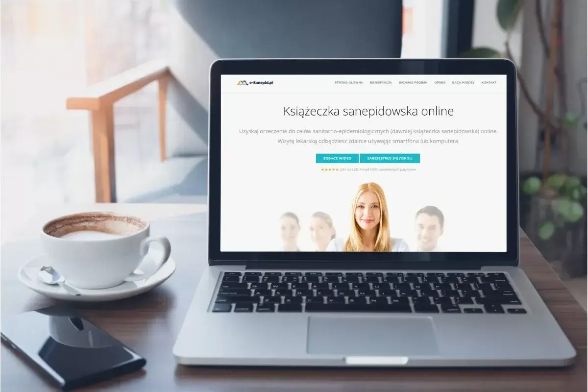 Laptop z kawą i telefonem. Na ekranie widnieje strona internetowa z ofertą "Książeczka sanepidowska online".