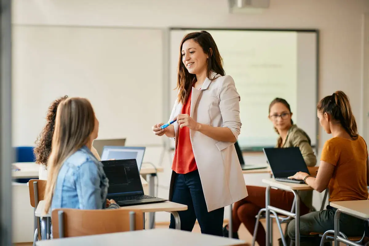 Nauczycielka wyjaśnia studentom, jak zostać nauczycielem zawodu. Uczniowie pracują na laptopach w sali lekcyjnej.