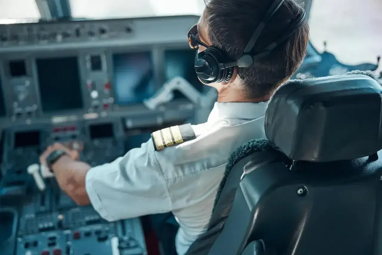 Pilot samolotu w słuchawkach i okularach przeciwsłonecznych, z ręką na sterach, w kabinie pełnej ekranów.