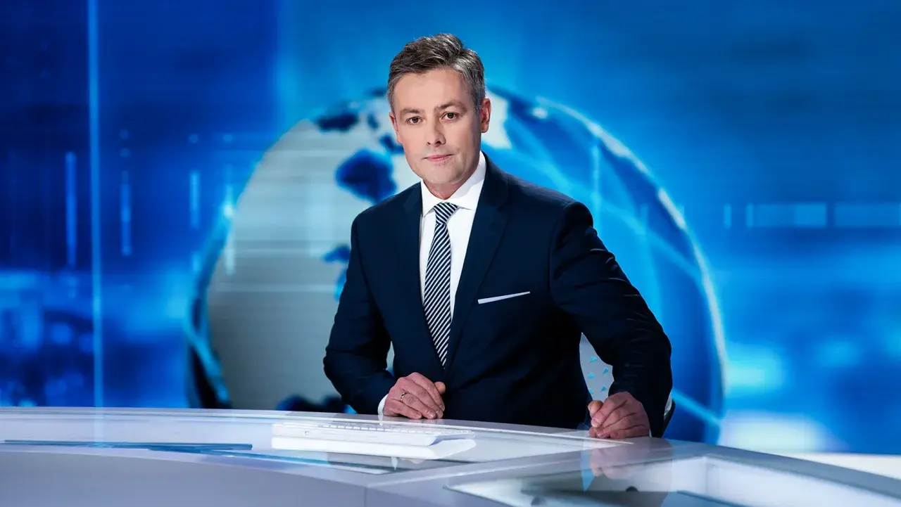 Elegancki konferansjer w garniturze stoi przy biurku na tle niebieskiej grafiki z globusem.