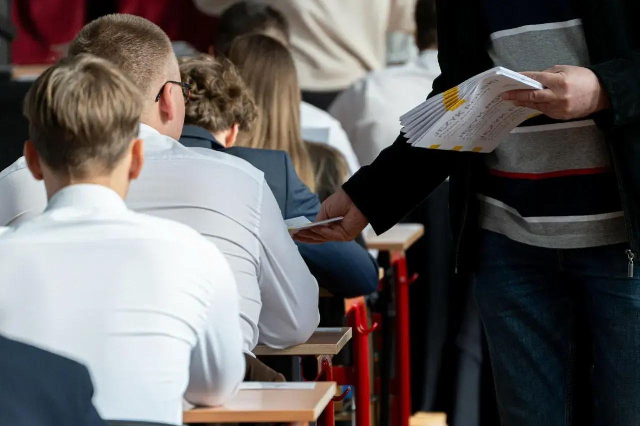 Uczniowie siedzą w ławkach, czekając na egzamin zawodowy sprzedawca. Nauczyciel rozdaje arkusze z zadaniami.