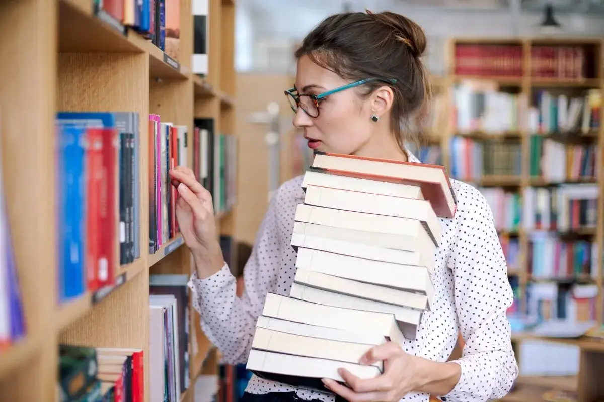 Młody bibliotekarz z okularami wybiera książki z półki, niosąc stos innych.