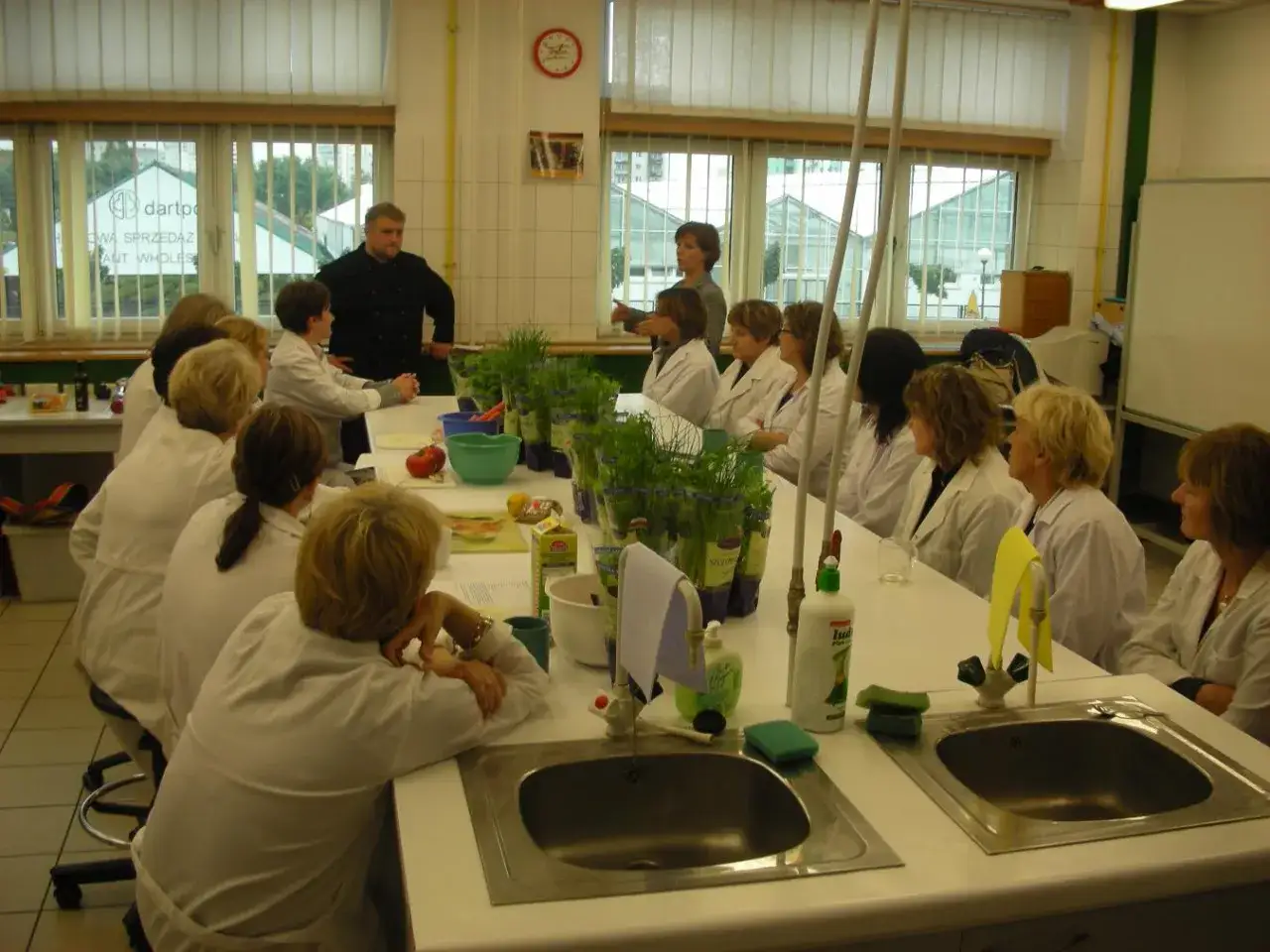 Intendentka prowadzi szkolenie dla grupy kobiet w białych fartuchach, prezentując zioła i warzywa na stole laboratoryjnym.
