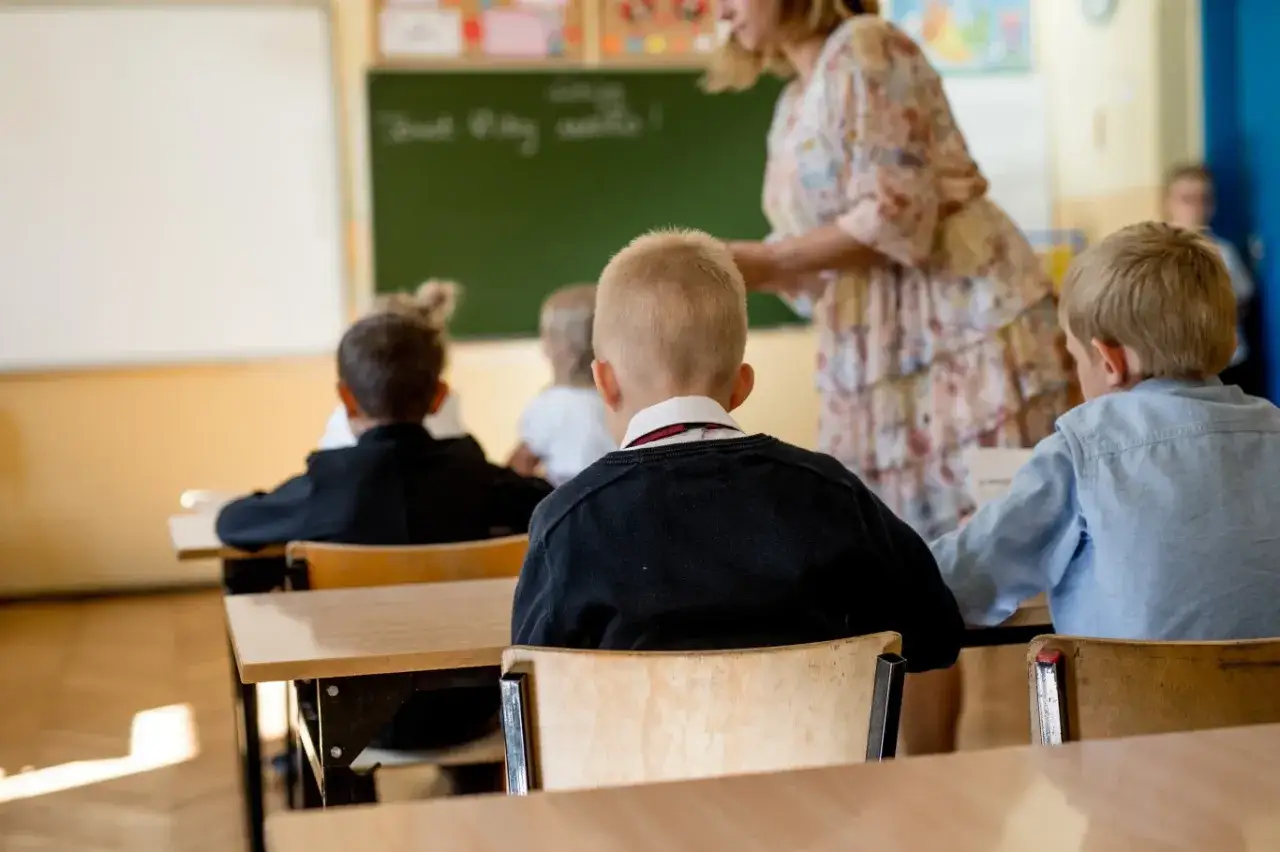 Uczniowie siedzą w ławkach, słuchając nauczycielki. Wszyscy czekają na swoje oceny z zachowania.
